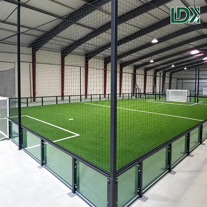 Glass football cage on an outdoor pitch, with lower glass panels, a tall mesh enclosure, goals and a green playing surface.